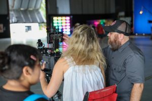 Three people work around a professional cinema camera on an indoor set. A bearded man in a black cap adjusts a monitor while a woman stands behind the camera, with colorful studio lights glowing in the background.