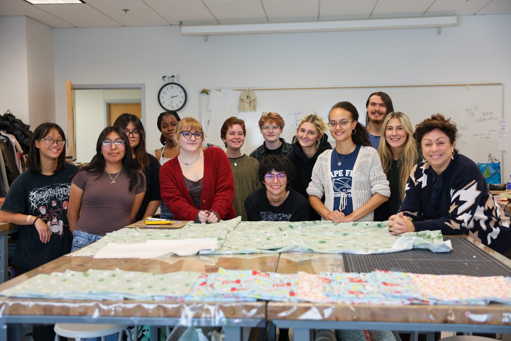 12 student members of the CFCC Sew Crafty Club and their advisor stand in front of a table covered in blankets that they have sewn to give to Novant for NICU babies.