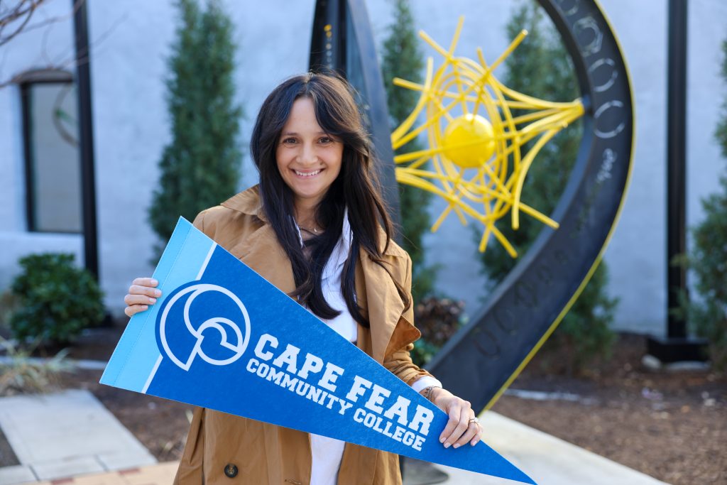 Smiling woman standing outdoors holding a blue Cape Fear Community College pennant, with a yellow abstract sculpture and greenery in the background. She is an author and Cape Fear Community College alumna.