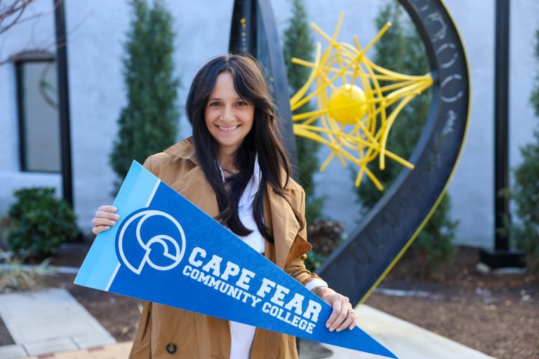 Smiling woman standing outdoors holding a blue Cape Fear Community College pennant, with a yellow abstract sculpture and greenery in the background. She is an author and Cape Fear Community College alumna.