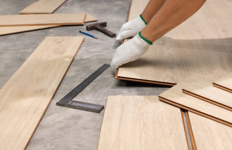 Hands wearing white work gloves align light wood flooring planks on a concrete floor, with a ruler, pencil, and mallet nearby.