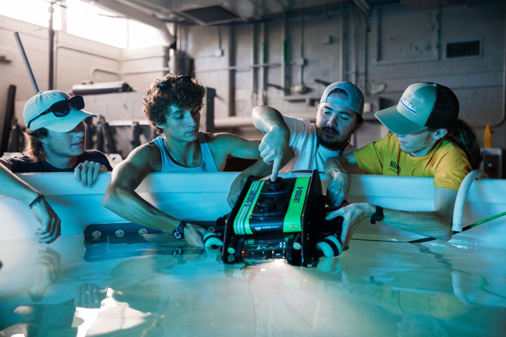 Four people lean over the edge of a large indoor water tank while adjusting a green and black underwater robotic device partially submerged in the water. One person points to a control knob on top of the device as the others hold it steady. The setting appears to be a workshop or lab with industrial equipment and pipes visible in the background.