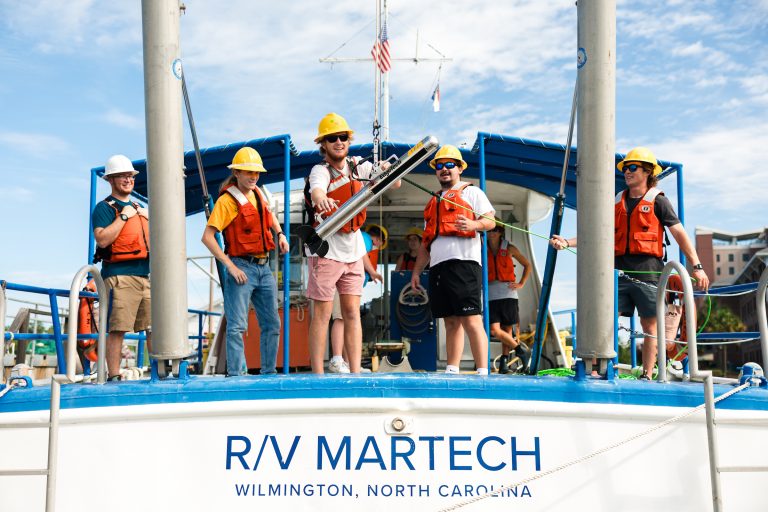 Here is ADA-compliant alt text: **Alt text:** Six students wearing hard hats and orange life vests stand on the deck of the research vessel R/V Martech in Wilmington, North Carolina, lowering a cylindrical oceanographic instrument into the water during a marine field exercise.