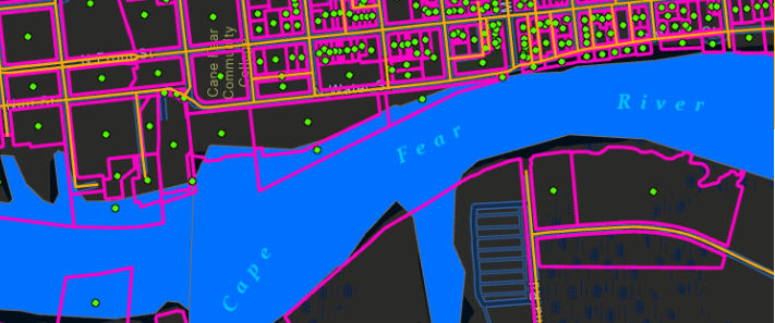 Color-coded map showing the Cape Fear River running horizontally through the center of the image. Surrounding the river are city blocks outlined in bright pink with a grid of streets highlighted in yellow. Numerous small green dots are scattered across the blocks, possibly marking specific locations. The labels “Cape Fear River” and “River” appear in light blue text over the water.