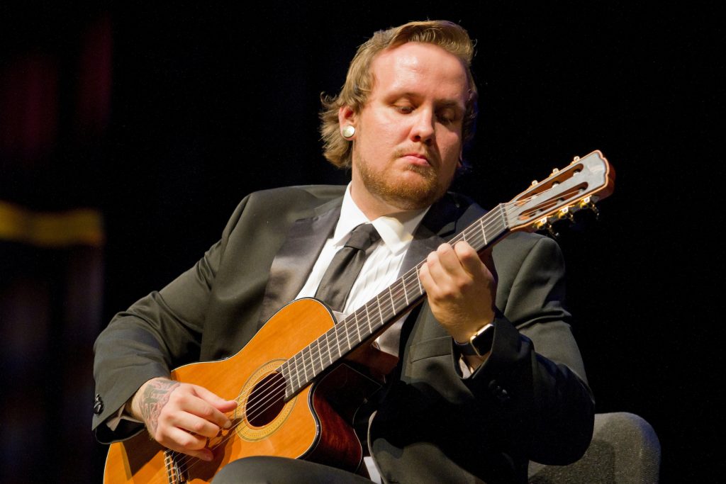 A musician in a suit sits onstage playing a classical acoustic guitar, focused on the instrument under stage lighting.
