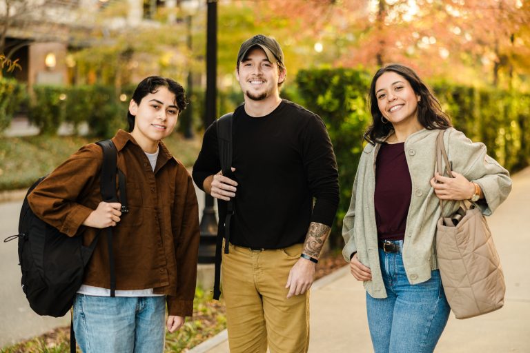 Three student veterans stand outdoors on a campus walkway in autumn, smiling at the camera. Each carries a bag or backpack, and trees with fall foliage and campus buildings are visible in the background.