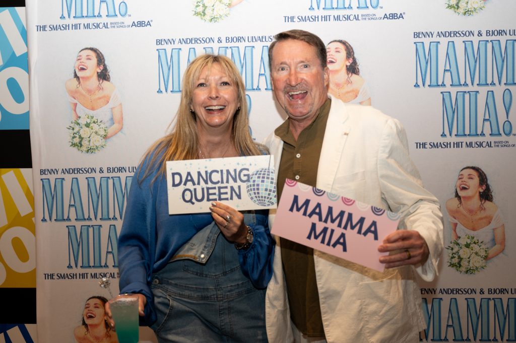Two adults smiling and posing in front of a step-and-repeat backdrop for the musical *Mamma Mia!*. The woman, wearing a blue sweater and denim overalls, holds a sign that reads “Dancing Queen” with a disco ball graphic and a blue-green drink in her other hand. The man, wearing a white jacket over a brown shirt, holds a pink sign that says “Mamma Mia.” The backdrop features repeated *Mamma Mia!* logos and images of a joyful bride holding a bouquet.