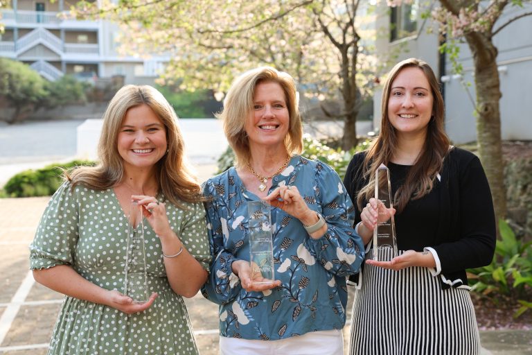 Three women stand outdoors on a sunlit walkway, smiling and facing the camera while holding clear glass award trophies. Behind them are blooming trees and a residential-style building with balconies, softly out of focus. The woman on the left wears a green dress with white polka dots, the woman in the center wears a blue patterned blouse, and the woman on the right wears a black top with a striped skirt.