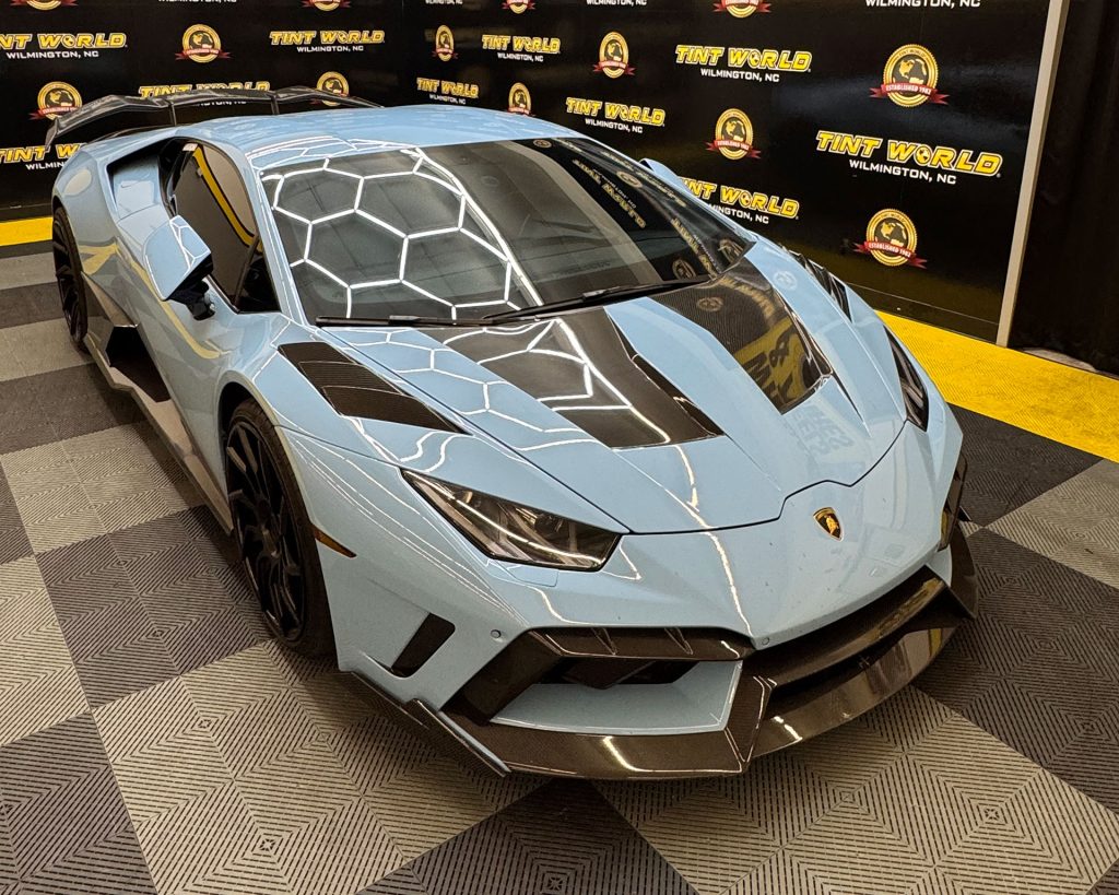 Light blue Lamborghini Huracán with black carbon-fiber hood accents and black wheels displayed indoors on a patterned showroom floor, photographed from the front-left angle. The background features a black promotional wall repeating the text “Tint World Wilmington, NC” with gold logos, and bright overhead lights reflect in a honeycomb pattern across the windshield and hood.
