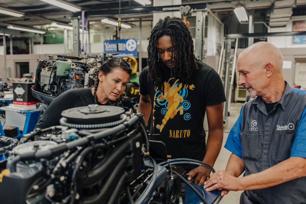 **Alt text:**
Three people stand in a workshop or technical training lab examining a large engine. An instructor in a work uniform points to a component while two students—a woman and a man—lean in and look closely at the machinery. Various mechanical equipment, tools, and fluorescent lights are visible in the background.