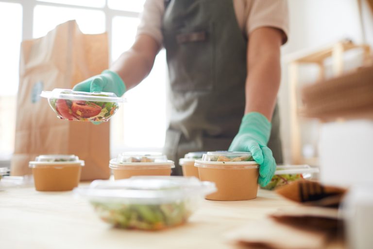 Food service worker wearing green gloves places a clear plastic container of salad on a table with other packaged meals, with a brown paper takeout bag visible in the background.