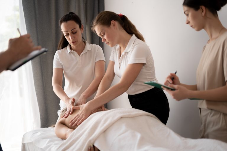 Three massage therapy trainees practice back-massage techniques on a client lying face down on a treatment table. Two women in white polo shirts use their hands to apply pressure to the client’s upper back while another woman stands nearby taking notes on a clipboard. The scene takes place in a bright treatment room with neutral walls, a curtain, and natural light coming from a window.