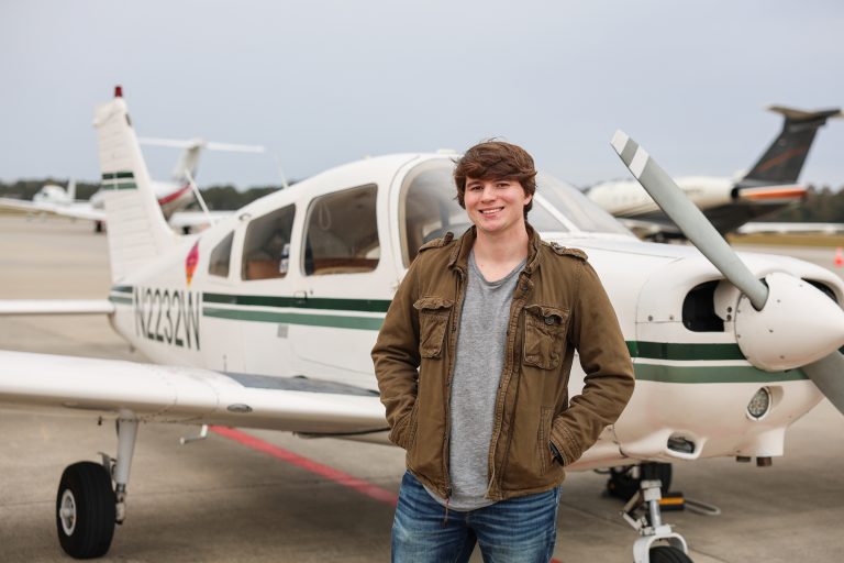 Young man standing in front of a small single-engine airplane on an airport tarmac, smiling with hands in his jacket pockets; other planes are visible in the background under an overcast sky.