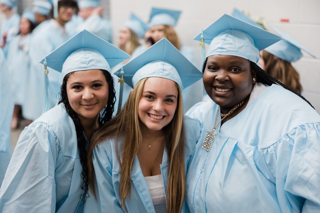 Three smiling graduates in light blue caps and gowns pose together in the foreground at a graduation ceremony, with other graduates blurred in the background.
