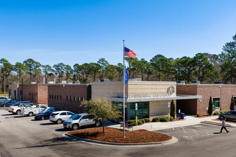 Wide exterior view of Cape Fear Community College's Surf City campus building on a clear day, with a U.S. flag and a blue college flag flying on a central flagpole. The modern, low-rise brick and beige building has the college name displayed above the entrance. Several cars are parked in a lot in front, landscaped shrubs and a small tree line the walkway, and a person walks near the entrance. Tall pine trees form a wooded backdrop behind the building.