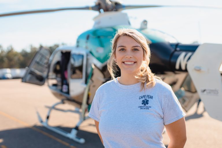 Young woman smiling and standing on an airport tarmac in front of a medical helicopter with its door open; she wears a light gray T-shirt that reads “Cape Fear Community College EMS Intern.