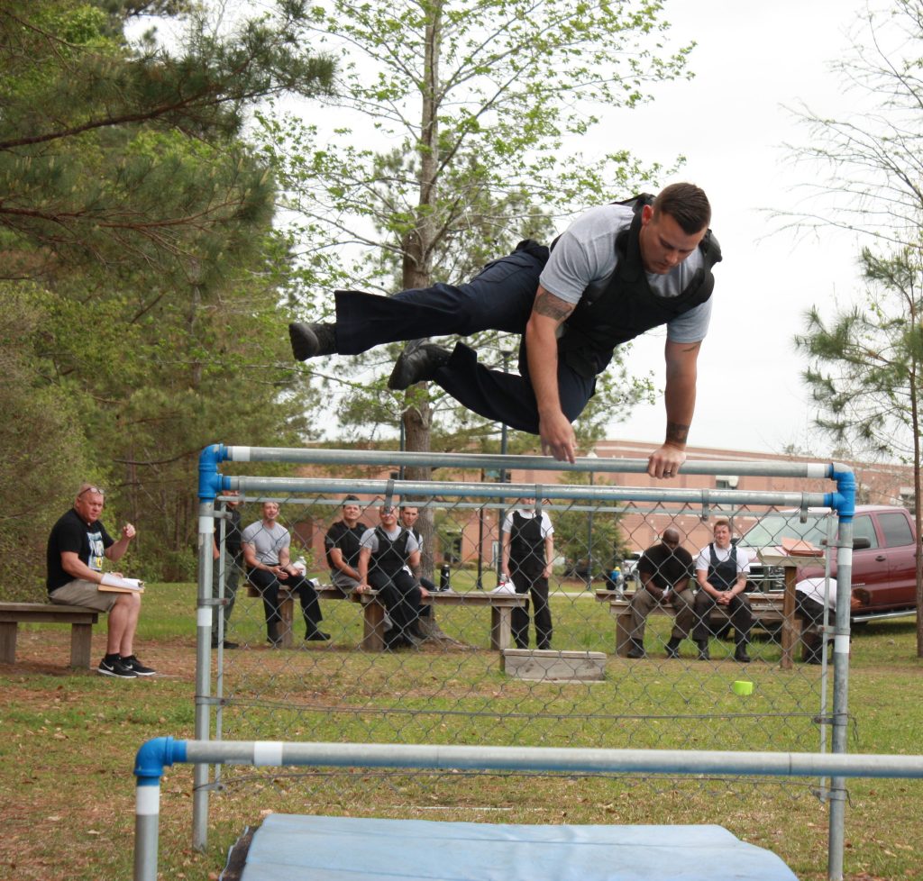 Man in athletic training attire vaults over a metal fence during an outdoor obstacle course, while a small group of onlookers in similar uniforms sit and watch from benches in a grassy area with trees and a parked vehicle in the background.