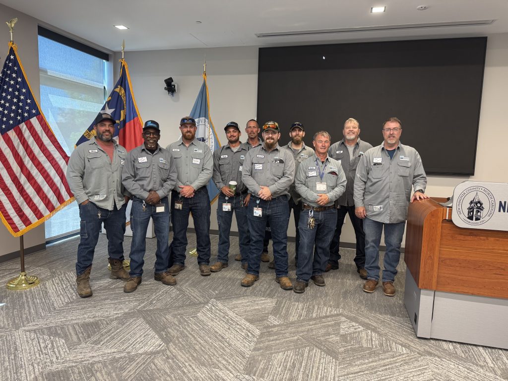 Alt text:
Group of approximately eleven adult men wearing matching gray work shirts, jeans, and work boots stand in a conference room, posing for a photo. They are arranged in two rows in front of a large blank presentation screen, with a wooden podium on the right displaying a county seal. Behind them are three flags, including the United States flag and a North Carolina state flag. Most of the men wear hats and ID badges, and several have beards. The room has large windows on the left and patterned carpet flooring.
