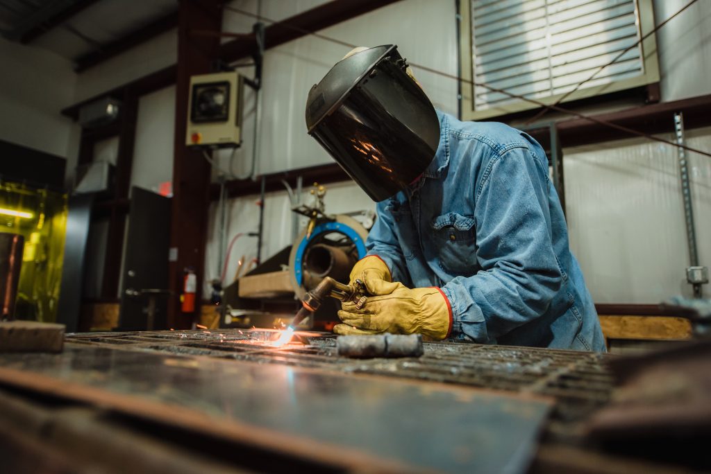 Worker wearing a welding helmet and heavy protective gloves uses a handheld torch to cut or weld metal on a workbench inside an industrial workshop, with sparks visible and machinery in the background.