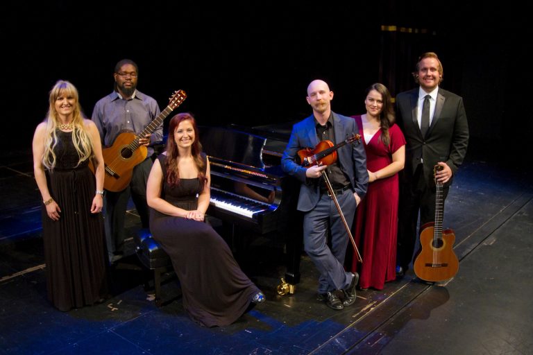 Group of six musicians posing on a stage around a grand piano; one person sits at the piano bench while others stand nearby holding instruments including guitars and a violin, dressed in formal and semi-formal attire against a dark theater background.