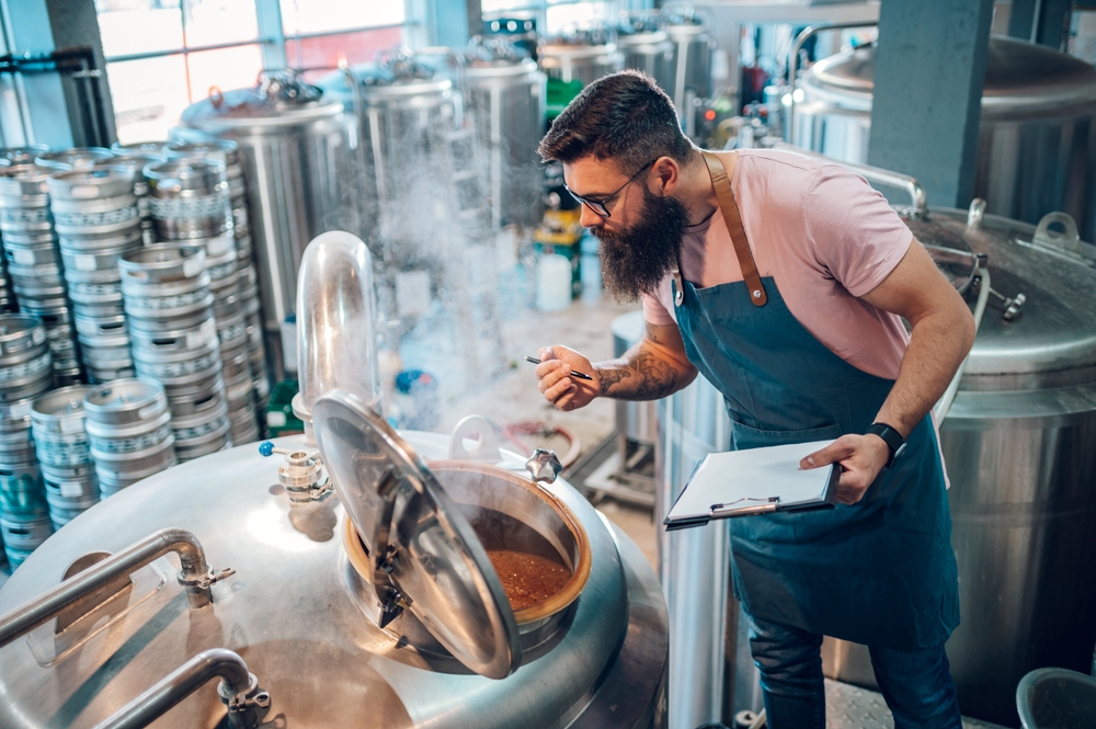 Bearded man wearing glasses and an apron inspects a steaming brewing tank, holding a clipboard and tool in a brewery filled with stainless steel equipment.