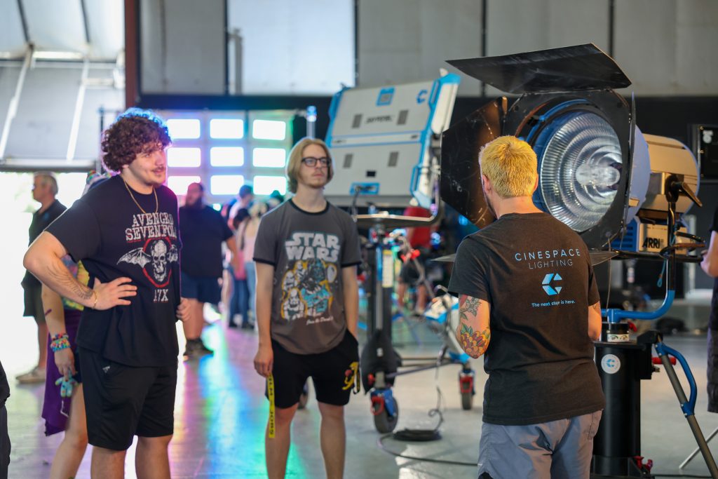 Three people stand in a film studio beside a large professional stage light while a crew member wearing a “Cinespace Lighting” shirt explains the equipment. Two young men in graphic T-shirts listen as colorful light from the background reflects across the polished floor, with additional lighting gear and people visible throughout the studio space.