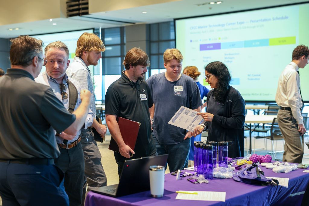 Group of adults gathered around a table at a professional event, engaged in conversation while examining printed materials. A woman on the right gestures to a sheet of paper as several men listen attentively. The table in the foreground is covered with a purple cloth and displays promotional items including reusable water bottles, small giveaways, and a laptop. In the background, large presentation screens show a schedule, and additional attendees stand and talk in a bright, modern conference room.

