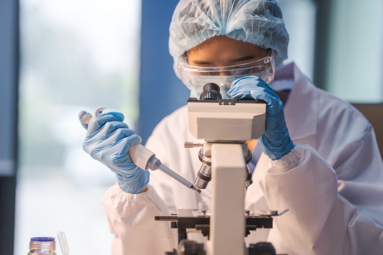 A laboratory researcher in protective gear uses a microscope while holding a pipette in a clinical lab setting. The person is wearing gloves, safety goggles, a face mask, and a hair cover while carefully handling a sample.