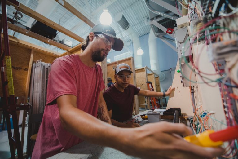 Two people wearing safety glasses work together in a technical training lab, examining and adjusting an electrical control panel with exposed wiring. The workspace contains tools, equipment, and partially constructed training stations in an industrial classroom setting.