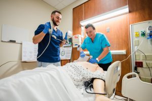 **ADA Alt Text:** Two nursing students in a clinical simulation lab practice emergency care on a medical training mannequin in a hospital bed. One student uses a bag-valve mask to provide ventilation while the other prepares a defibrillator. The room includes patient monitoring equipment, wall-mounted medical gas outlets, and clinical training materials. 