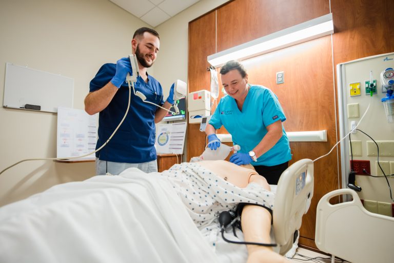 **ADA Alt Text:** Two nursing students in a clinical simulation lab practice emergency care on a medical training mannequin in a hospital bed. One student uses a bag-valve mask to provide ventilation while the other prepares a defibrillator. The room includes patient monitoring equipment, wall-mounted medical gas outlets, and clinical training materials.