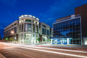 **ADA Alt Text:** Nighttime exterior view of Union Station and the adjacent Sea Devil Bookstore, featuring modern multi-story buildings with illuminated windows and glass façades. Long-exposure light trails from passing vehicles streak across the street in the foreground beneath a deep blue evening sky.