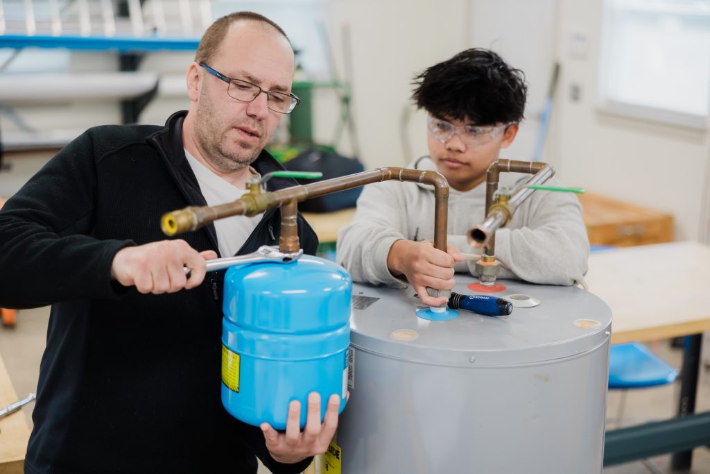 Instructor and student wearing safety glasses work together on a water heater training project, connecting copper pipes and components in a classroom workshop.