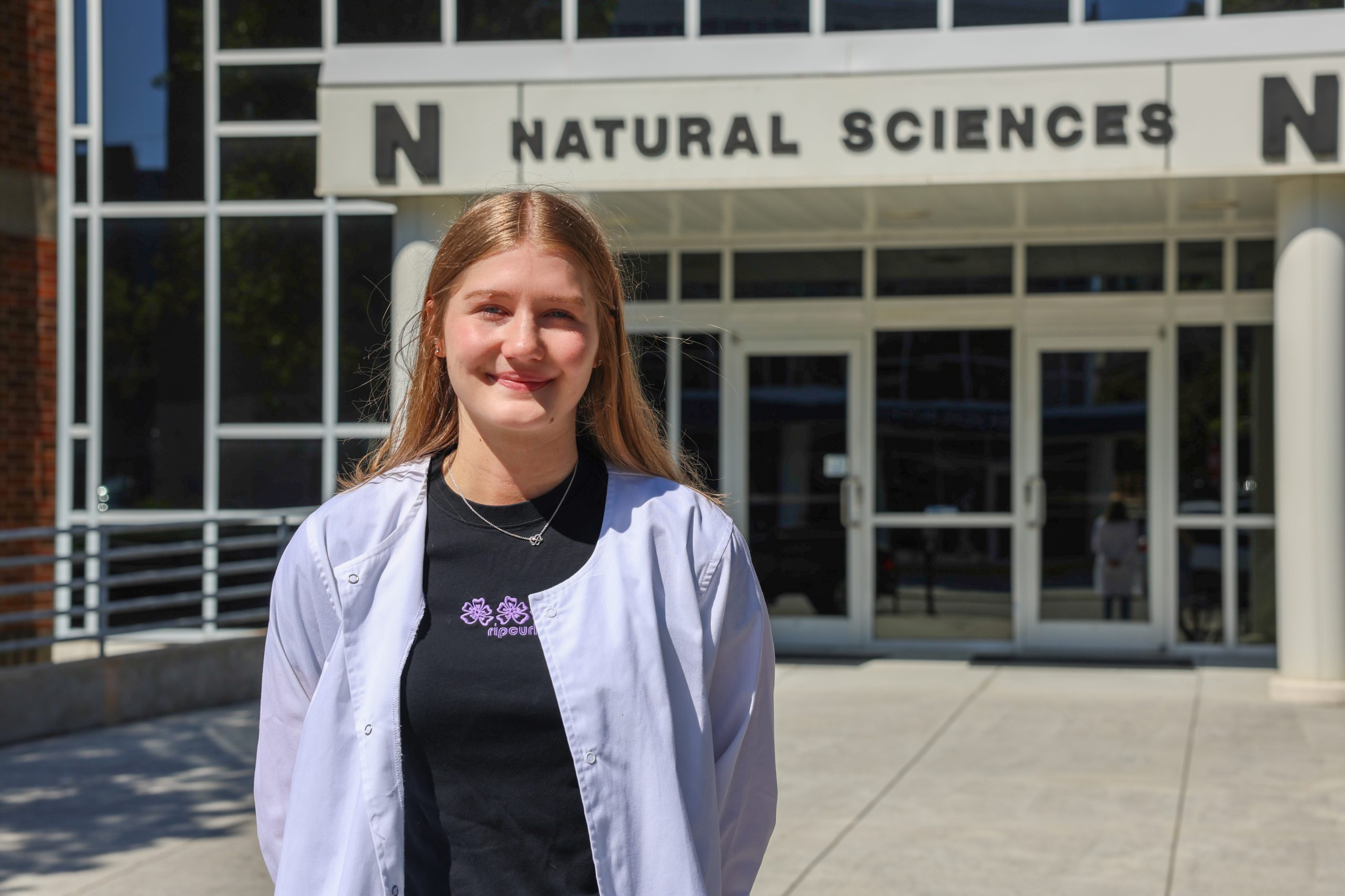 student in white lab coat standing outside Natural Sciences building