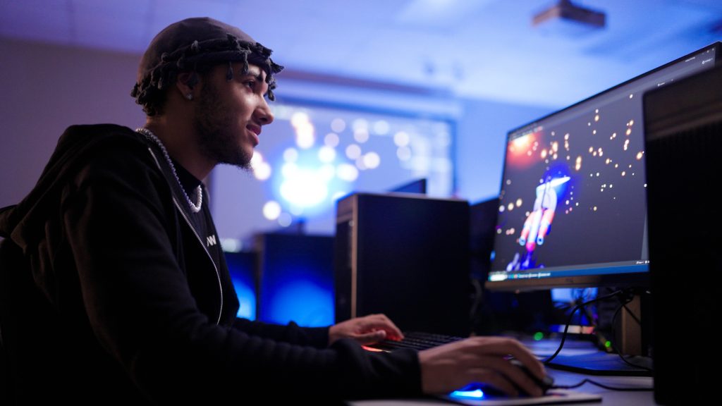 A person wearing a dark hoodie and head covering sits at a desktop computer in a dimly lit room with blue and purple ambient lighting. They are using a keyboard and mouse while looking at a large monitor displaying a colorful 3D character or animation. Several computer towers and monitors are visible around the workstation, creating a technology-focused environment.
