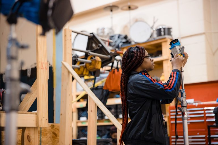 A person with long braided hair adjusts a studio light mounted on a stand in a production workshop. Shelving, equipment, and a red tool chest are visible in the background, suggesting a film or photography studio environment.