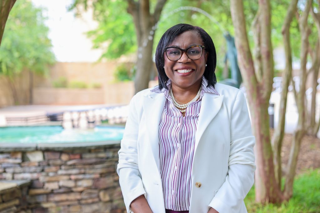 Smiling woman in a white blazer standing outdoors near a fountain and trees.