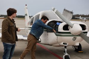 Two men stand beside a small propeller airplane on an airport tarmac while one reaches into the aircraft’s open engine compartment.