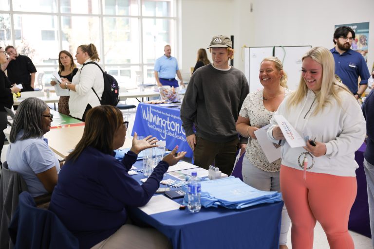 tudents and community members talk with representatives at information tables during a busy indoor resource fair. In the foreground, two smiling attendees holding papers speak with a seated representative, while other groups mingle near tables in the bright, window-filled room