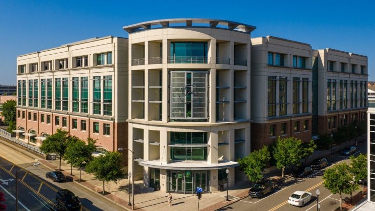 Modern multi-story institutional building with a curved central entrance, large green-tinted windows, and brick-and-concrete exterior, viewed from above at a city street intersection on a clear day.