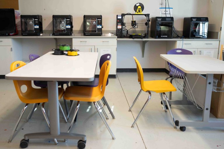 Classroom makerspace with several 3D printers arranged on countertops, colorful filament spools, white worktables, and yellow and purple chairs.