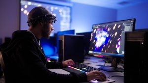 A person sits at a desktop computer in a dimly lit computer lab, using a keyboard and mouse while focusing on a widescreen monitor displaying a colorful digital graphics or animation program. Multiple computer towers and monitors surround the workstation, with blue ambient lighting creating a modern technology workspace.