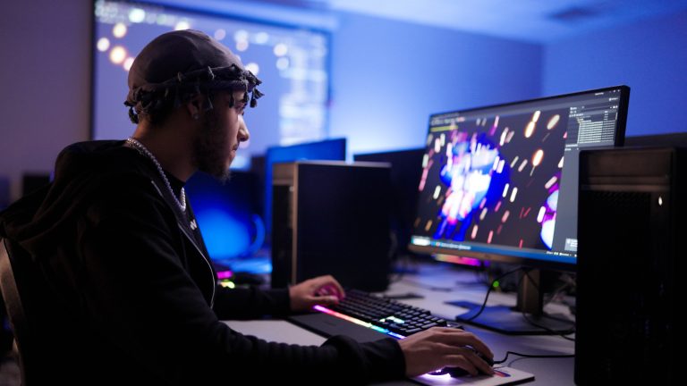 A person sits at a desktop computer in a dimly lit computer lab, using a keyboard and mouse while focusing on a widescreen monitor displaying a colorful digital graphics or animation program. Multiple computer towers and monitors surround the workstation, with blue ambient lighting creating a modern technology workspace.