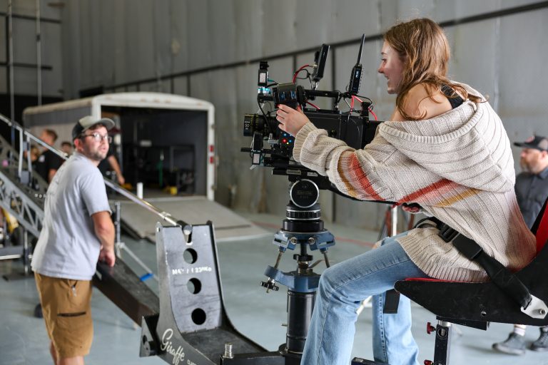 A camera operator sits on a mounted seat while adjusting a large professional cinema camera on a studio set. Crew members, production equipment, and a trailer are visible in the background.
