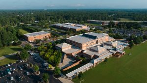 Aerial view of a large brick school campus with multiple connected buildings, parking lots, covered walkways, athletic fields, and surrounding trees.