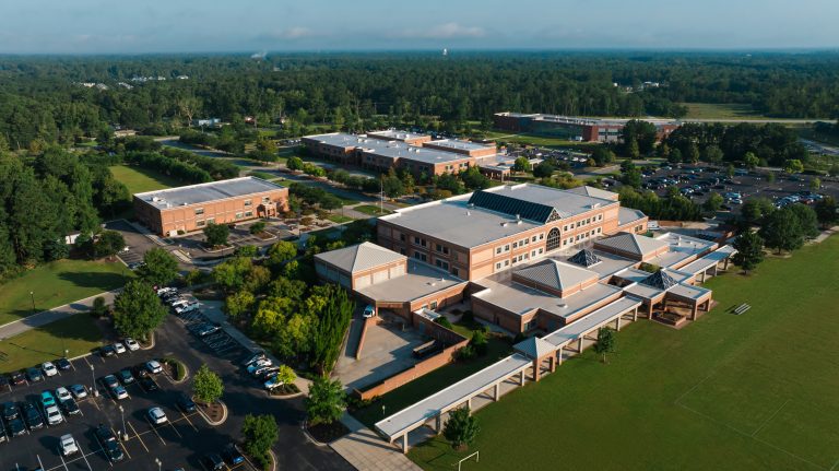 Aerial view of a large brick school campus with multiple connected buildings, parking lots, covered walkways, athletic fields, and surrounding trees.