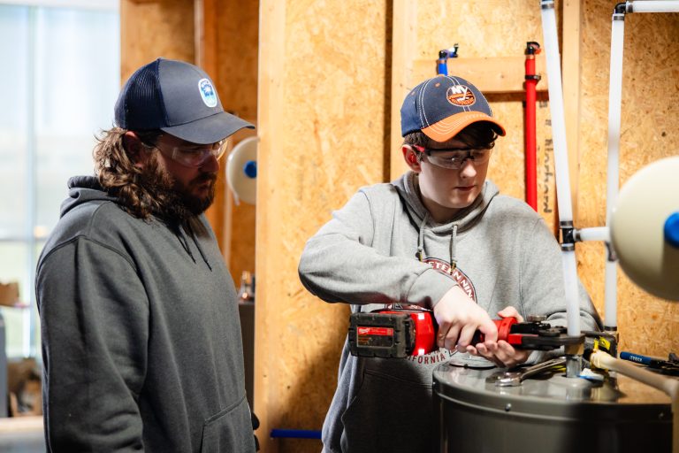 A student wearing safety glasses uses a red power tool to work on piping connected to a large tank while another man observes in a workshop.