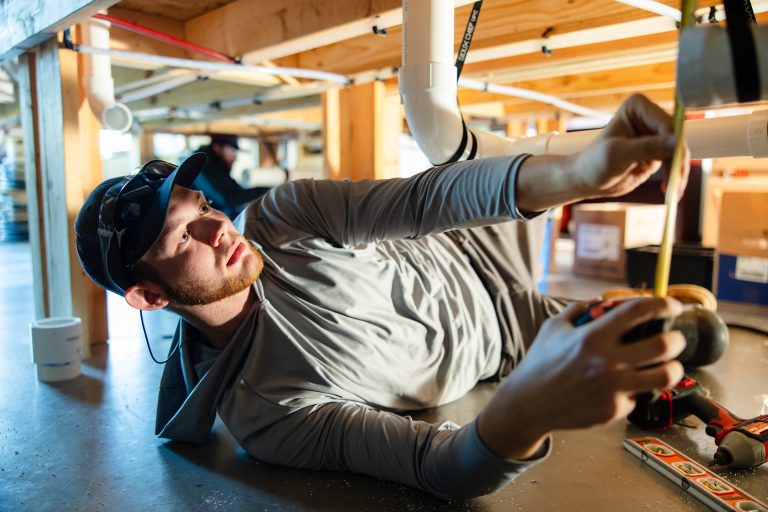 A construction worker wearing a cap and safety glasses lies beneath exposed plumbing, reaching upward while using a power tool during installation work.