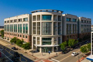 Modern multi-story institutional building with a curved central entrance, large green-tinted windows, and brick-and-concrete exterior, viewed from above at a city street intersection on a clear day.