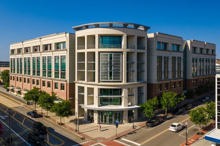 Modern multi-story institutional building with a curved central entrance, large green-tinted windows, and brick-and-concrete exterior, viewed from above at a city street intersection on a clear day.